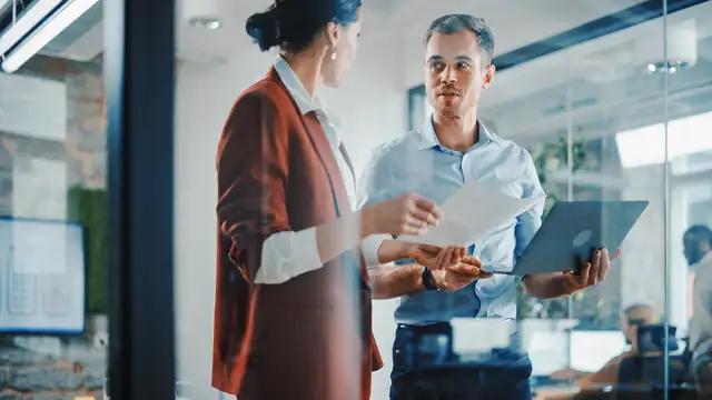 Woman and man holding a paper and computer discussing something in a office